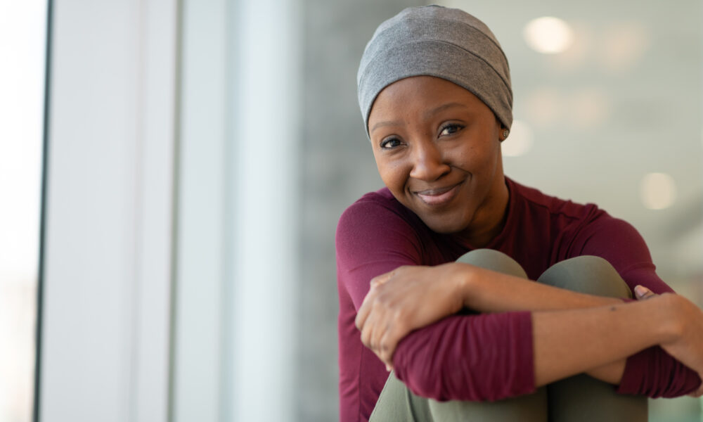 A beautiful woman with alopecia wears a hat. The woman is smiling at the camera.